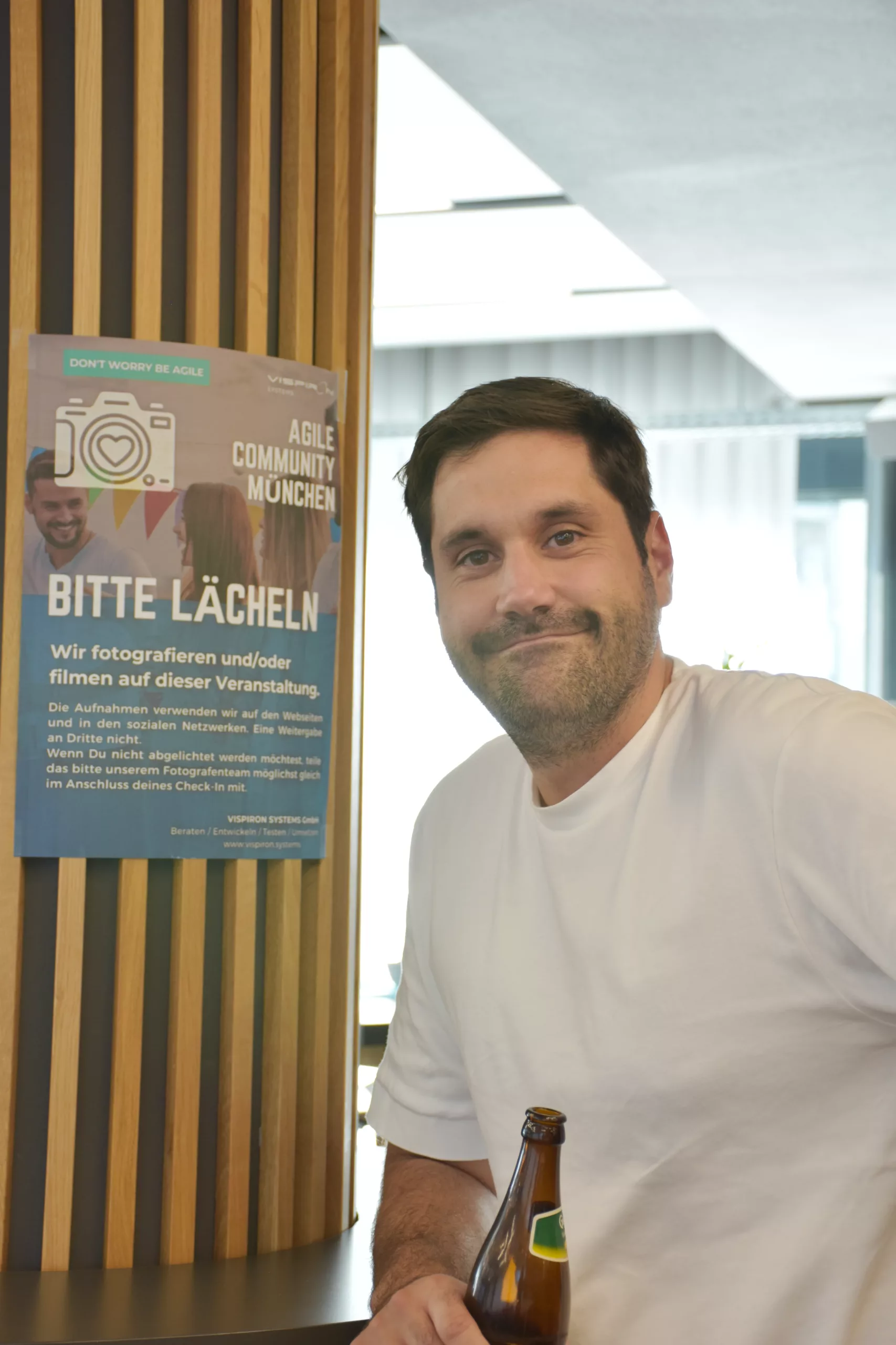 Foto eines Teilnehmenden der Agile Community München vor einem Plakat Bitte lächeln